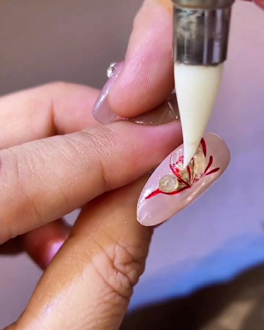 Un outil en silicone pointu pressant et aplatissant une perle de gel de modelage transparent sur une aile de papillon rouge et or.