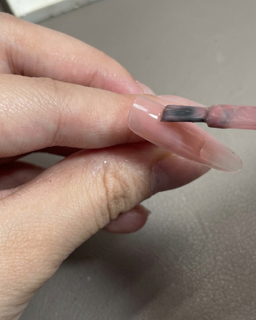 Gros plan sur un pinceau appliquant un vernis gel rose translucide sur une capsule dongle claire.