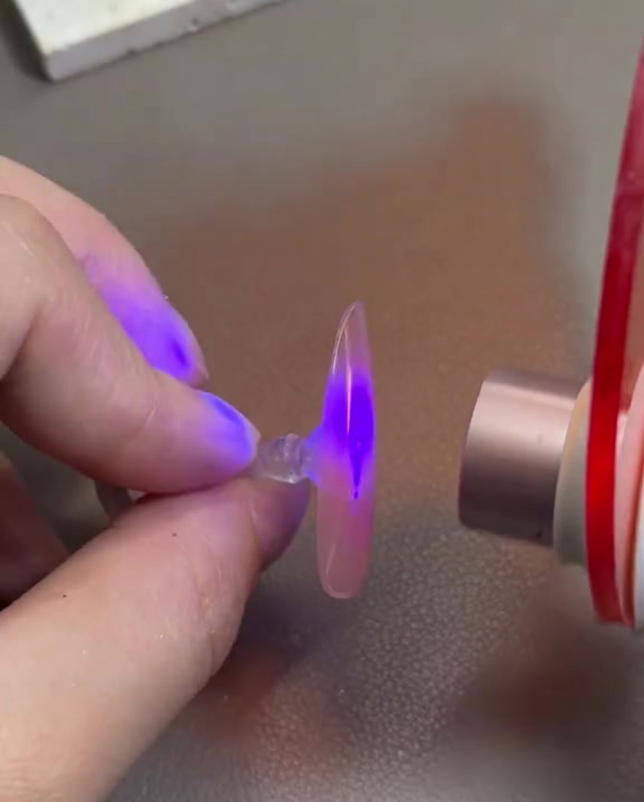 Une main tenant une capsule dongle avec du gel rose sous une mini lampe de catalyse UV/LED.