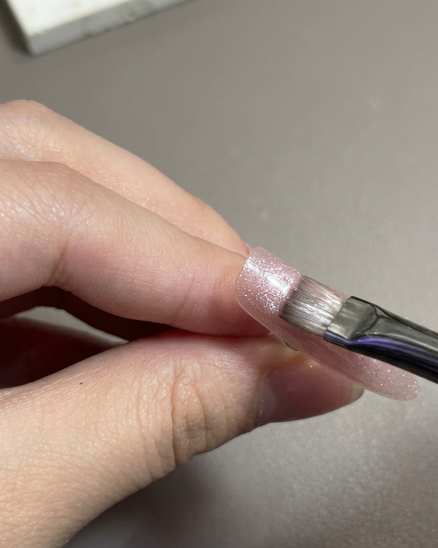 Un pinceau plat à gel étalant du gel magnétique œil de chat rose scintillant sur une capsule dongle.