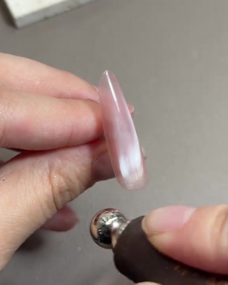 Une main tenant un outil aimanté cylindrique près dun ongle long peint avec du gel œil de chat rose humide, déplaçant les particules magnétiques.