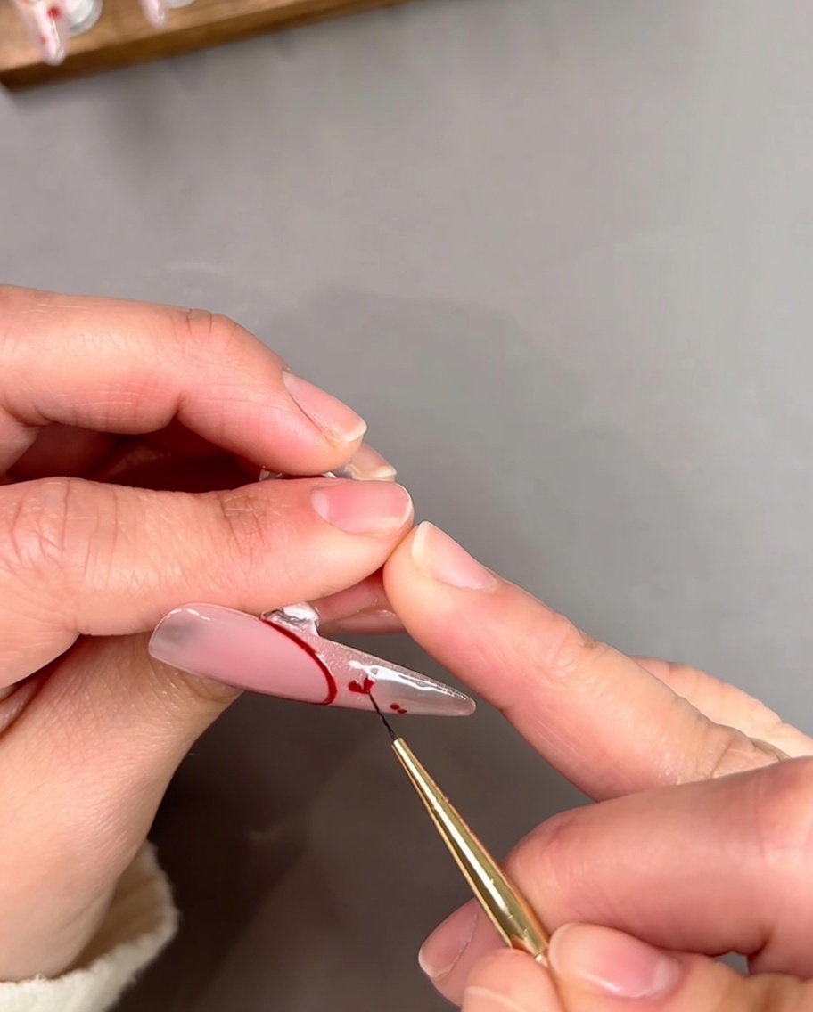 Gros plan dune styliste ongulaire utilisant un pinceau fin pour relier deux petits points rouges en forme de cœur sur un embout dongle transparent.
