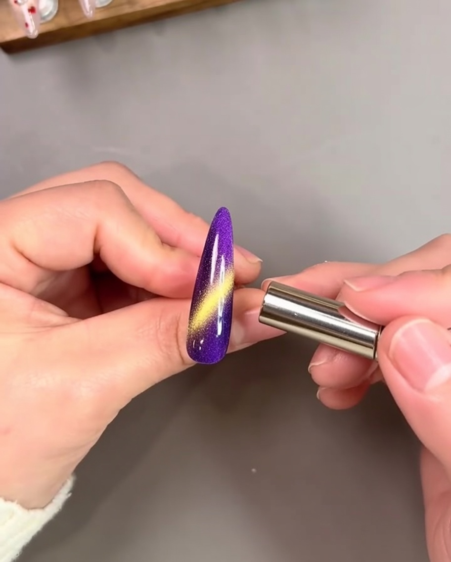 Une main tenant un aimant cylindrique près dun ongle peint en violet vibrant, activant une traînée œil-de-chat dorée brillante.