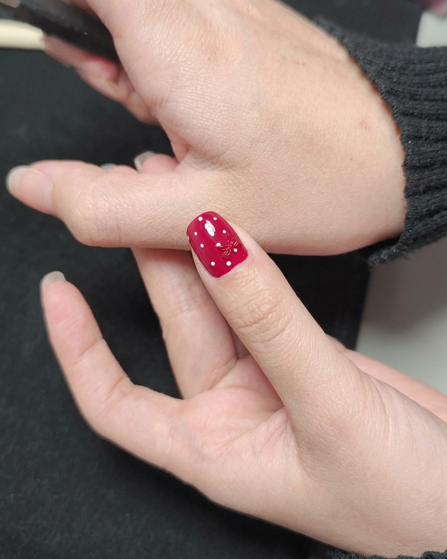 Un pinceau à vernis appliquant un top coat transparent brillant sur un ongle rouge avec des pois blancs et un nœud rouge.