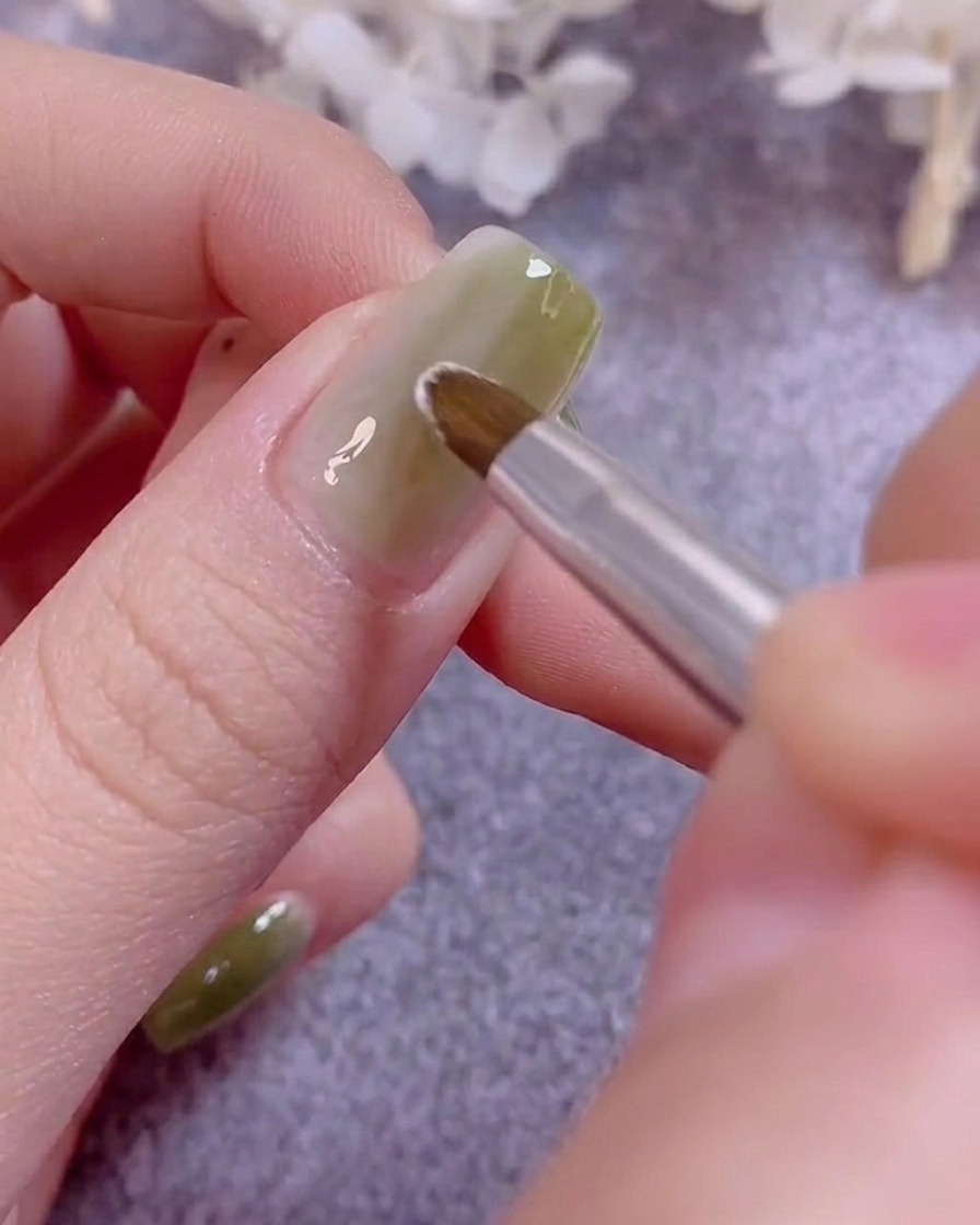 Un pinceau de détail fin peignant de délicats pétales de fleurs blancs sur un ongle recouvert de gel vert.
