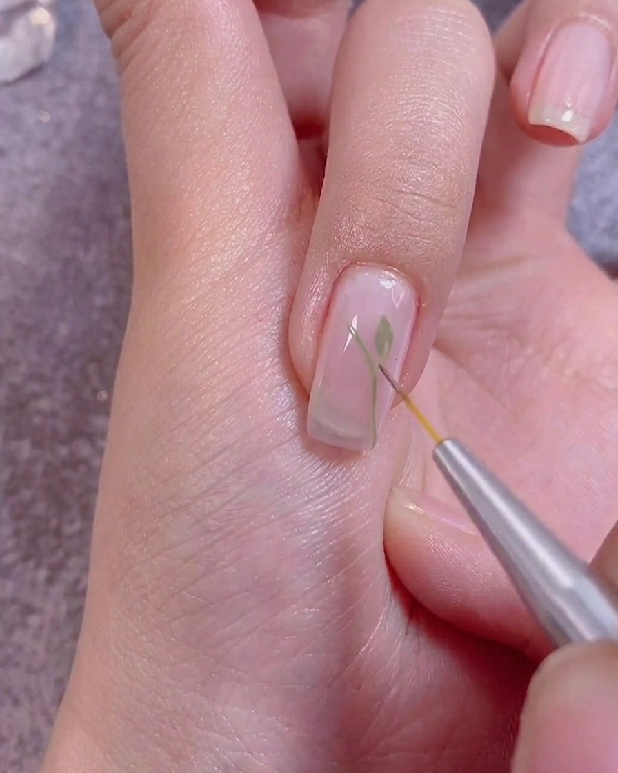Gros plan montrant un pinceau fin ajoutant des détails blancs sur des feuilles vertes peintes sur un ongle.