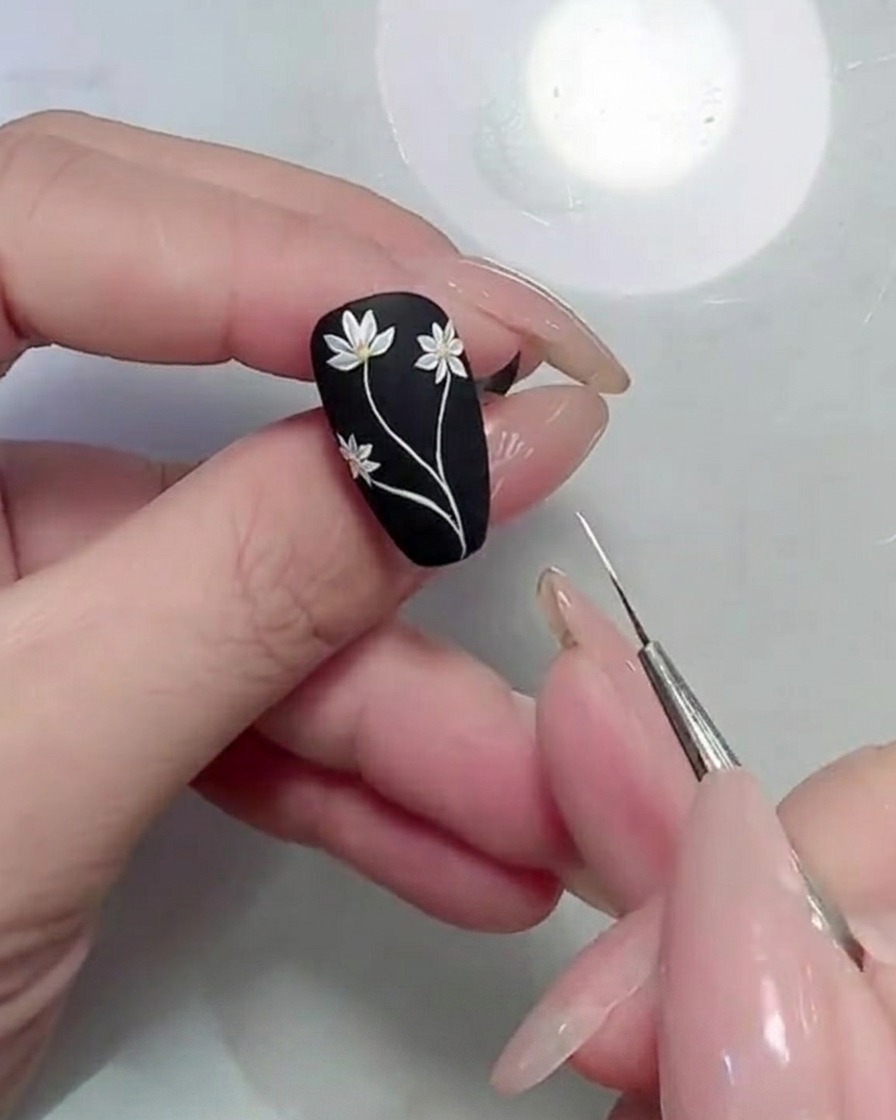 Gros plan dun pinceau détail appliquant des formes de feuilles vertes près des tiges sur un ongle noir mat.
