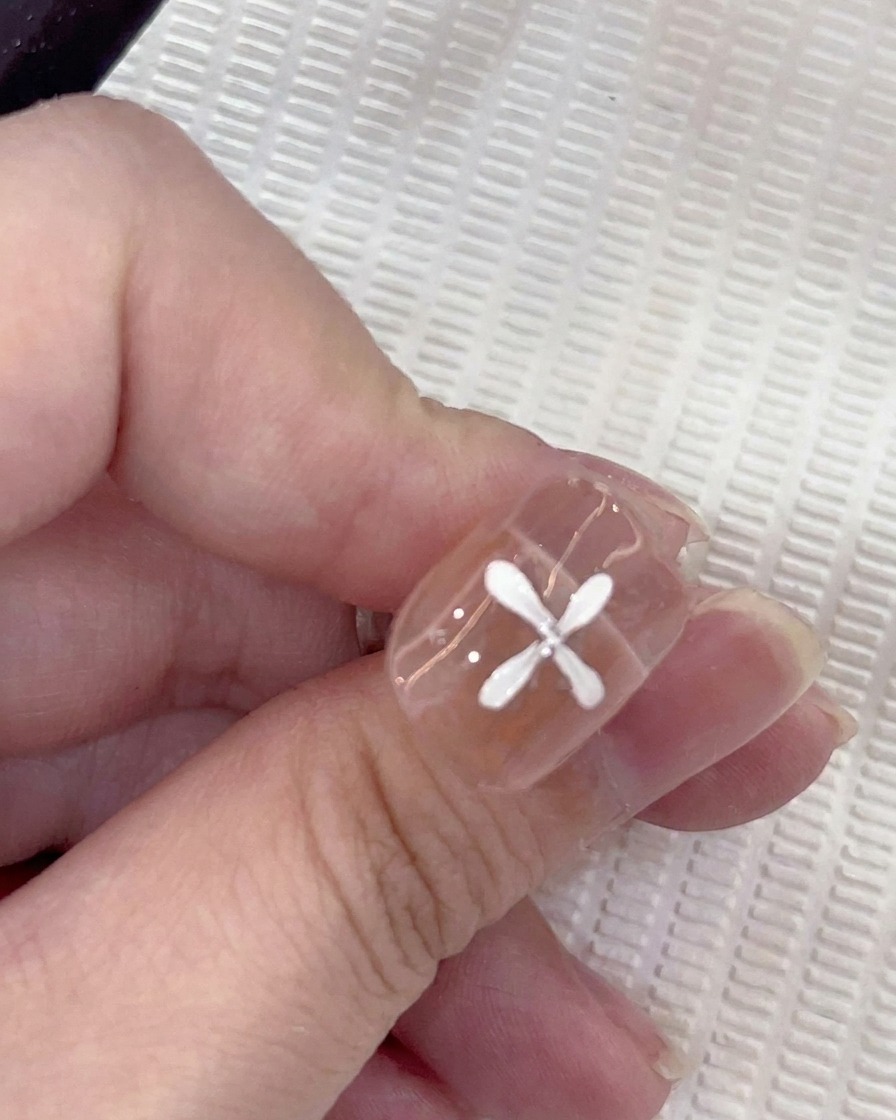 Gros plan dun ongle avec cinq pétales blancs peints en motif circulaire, avec un pinceau ajoutant un sixième pétale.