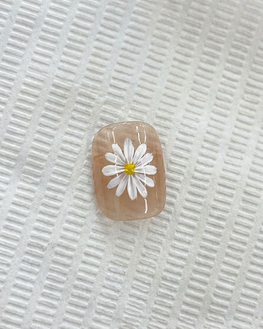 Gros plan dune marguerite blanche sur un ongle, avec un pinceau de détail fin appliquant un centre jaune.