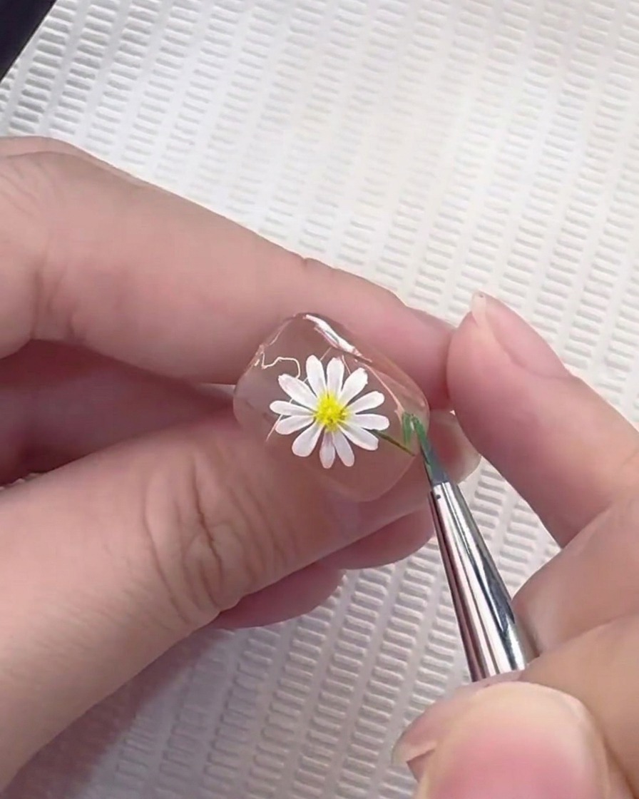 Un pinceau liner tirant une fine ligne verte pour former une tige sur un ongle décoré de marguerites.