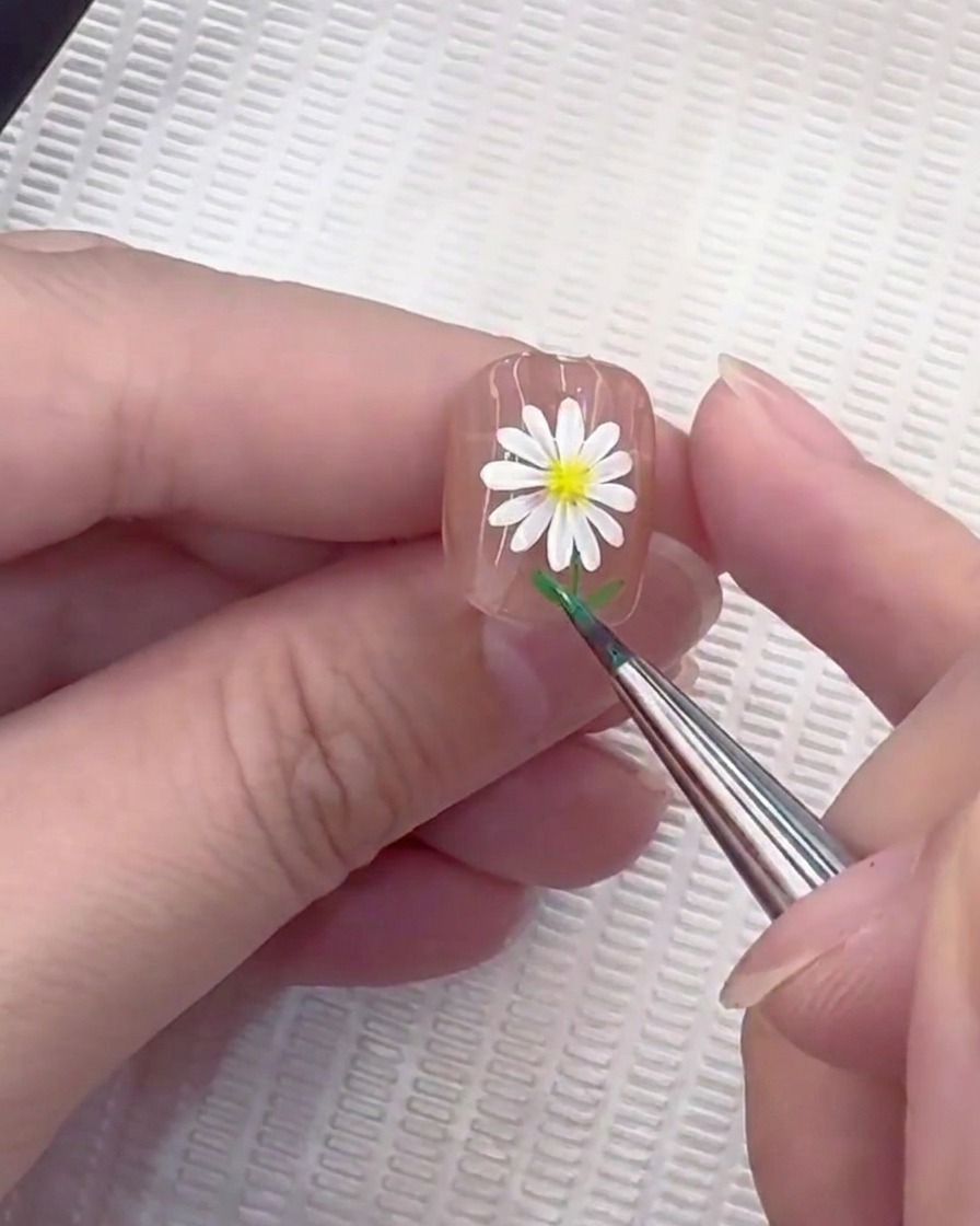 Un pinceau liner ajoutant de petites feuilles vertes à la base dune tige peinte sur un ongle.