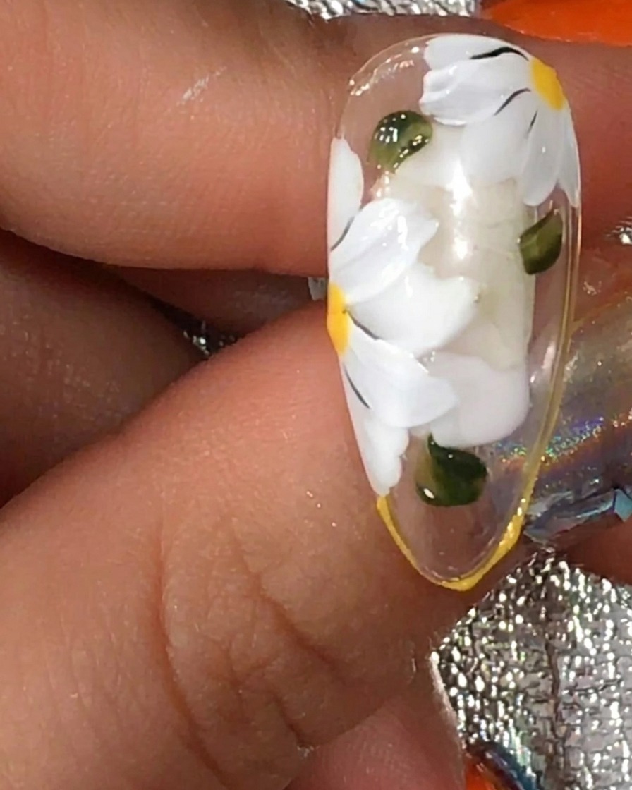 Un pinceau appliquant une couche finale de top coat mat sur un ongle avec un design de marguerite blanche et feuilles vertes.