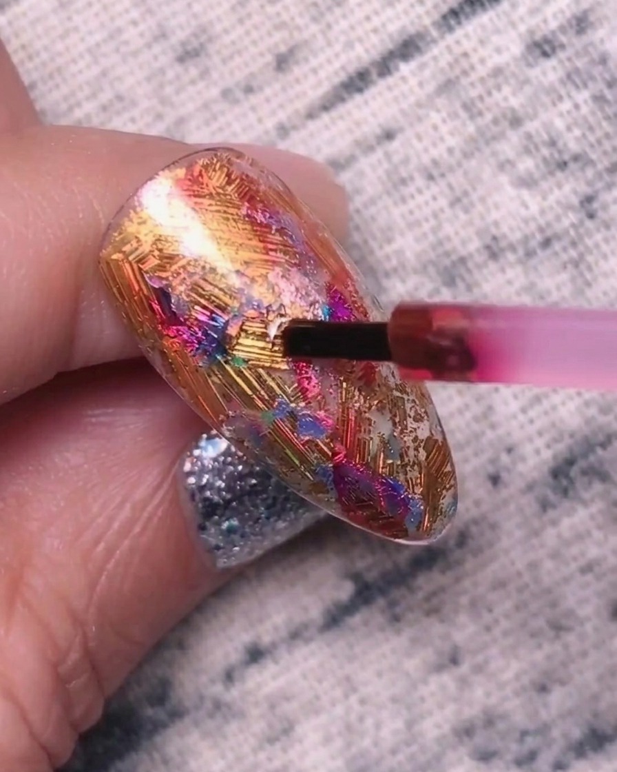 Un petit pinceau appliquant un gel pigmenté rouge-violet sur une surface dongle texturée recouverte de feuilles métalliques.