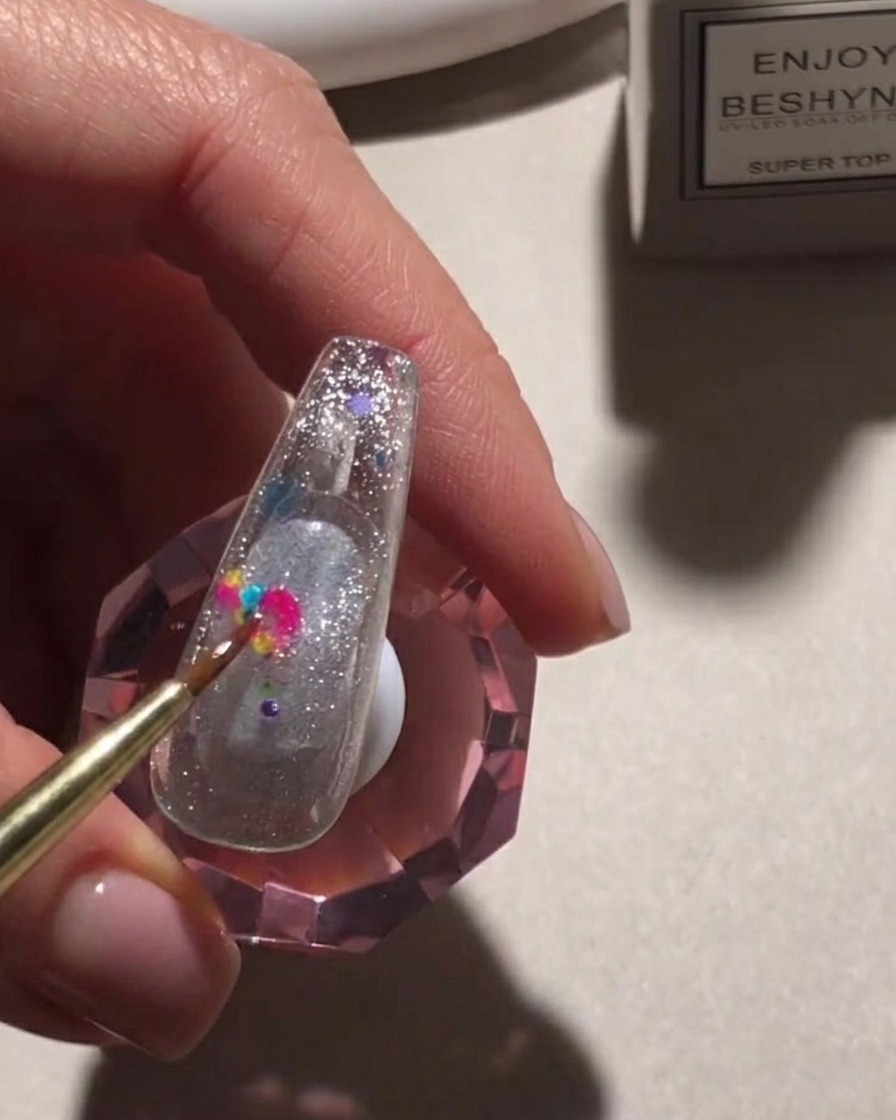 Un pinceau de précision déposant des sequins ronds multicolores sur une surface dongle pailletée.