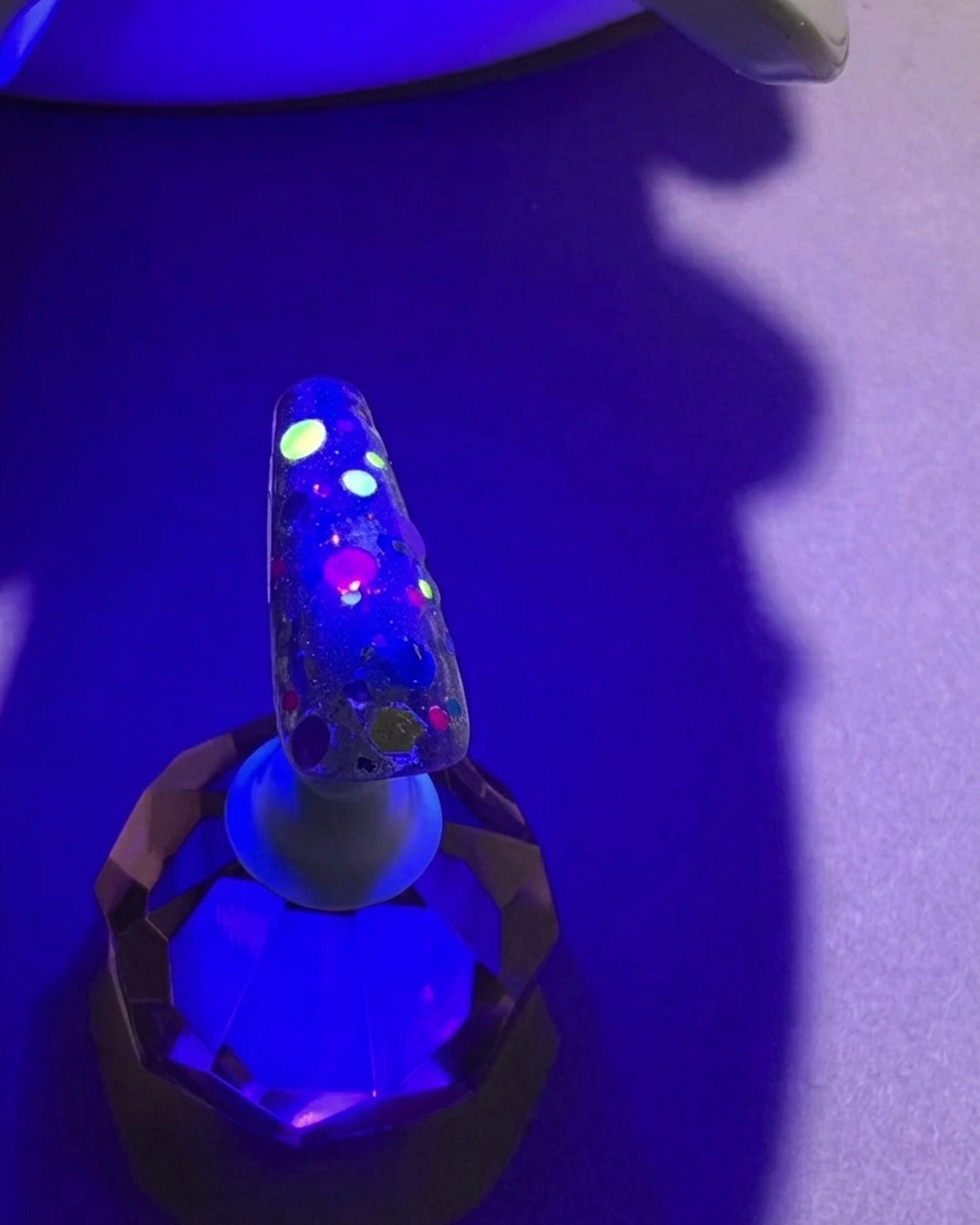 Un ongle avec des sequins colorés sous la lumière violette dune lampe UV, pendant la catalysation.