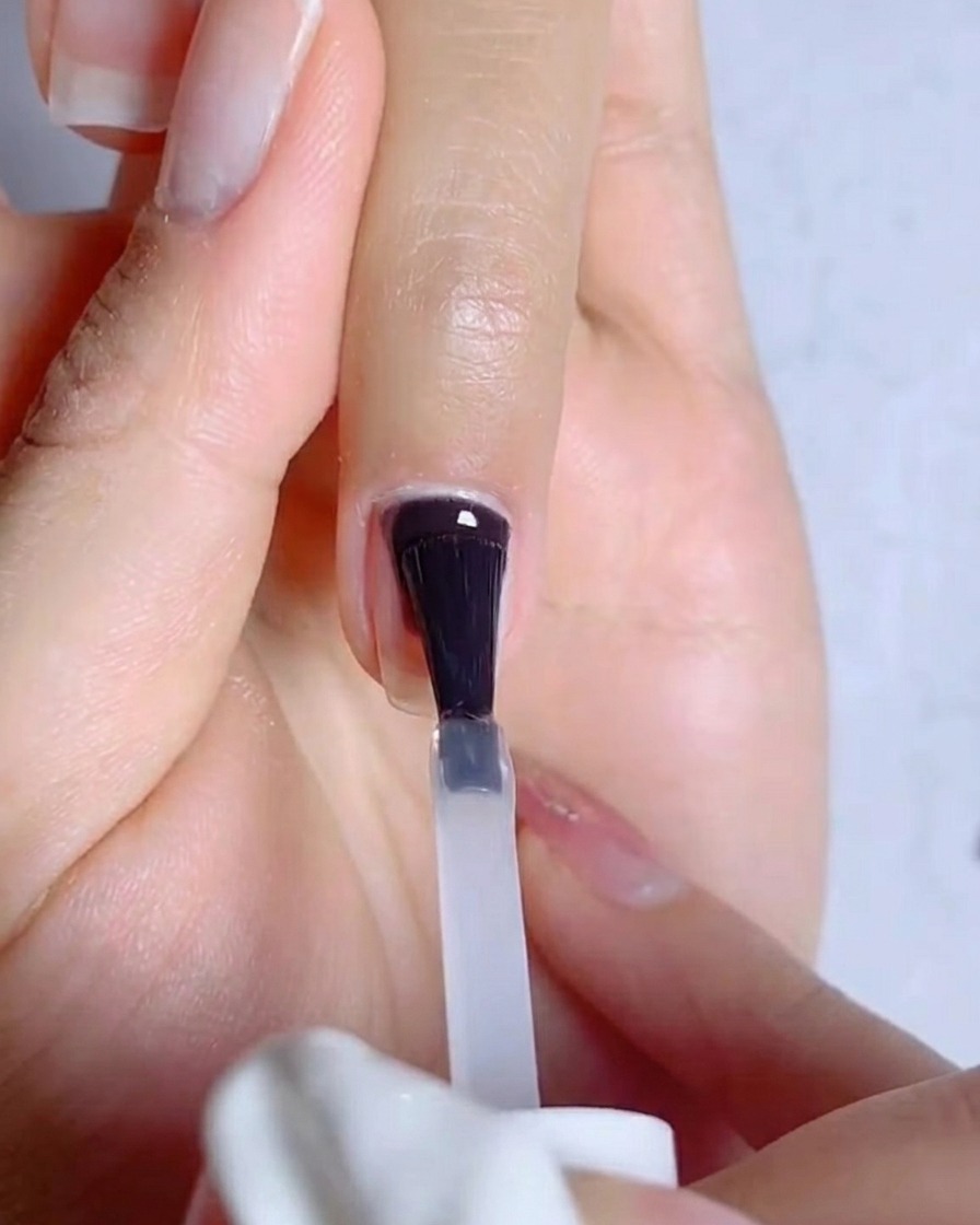 Un gros plan montrant un pinceau appliquant une fine couche transparente de base gel sur un ongle naturel préparé.