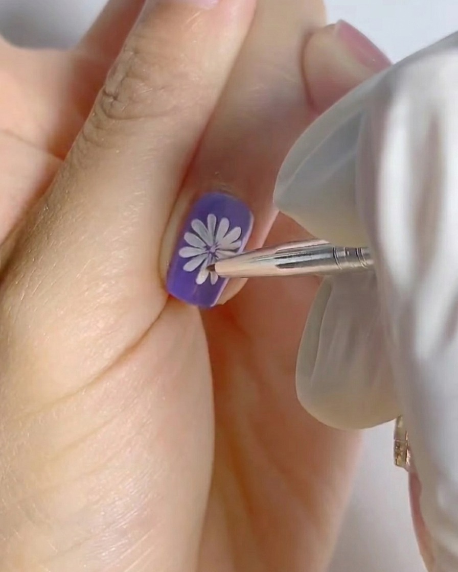 Un doigt avec le nail art marguerite blanche terminé sur base violette placé sous une petite lampe professionnelle pour la catalyse.