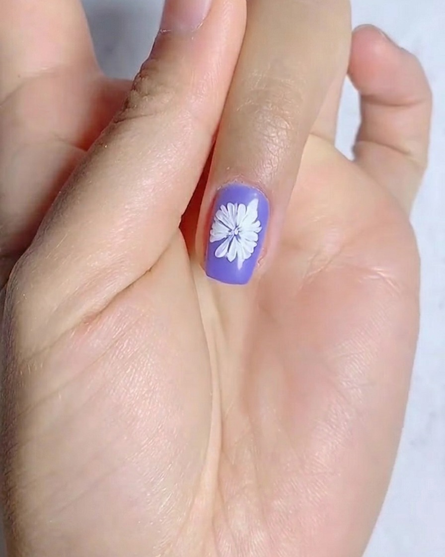 Une vue rapprochée montrant un point jaune étant appliqué au centre dune marguerite blanche peinte sur un ongle violet.