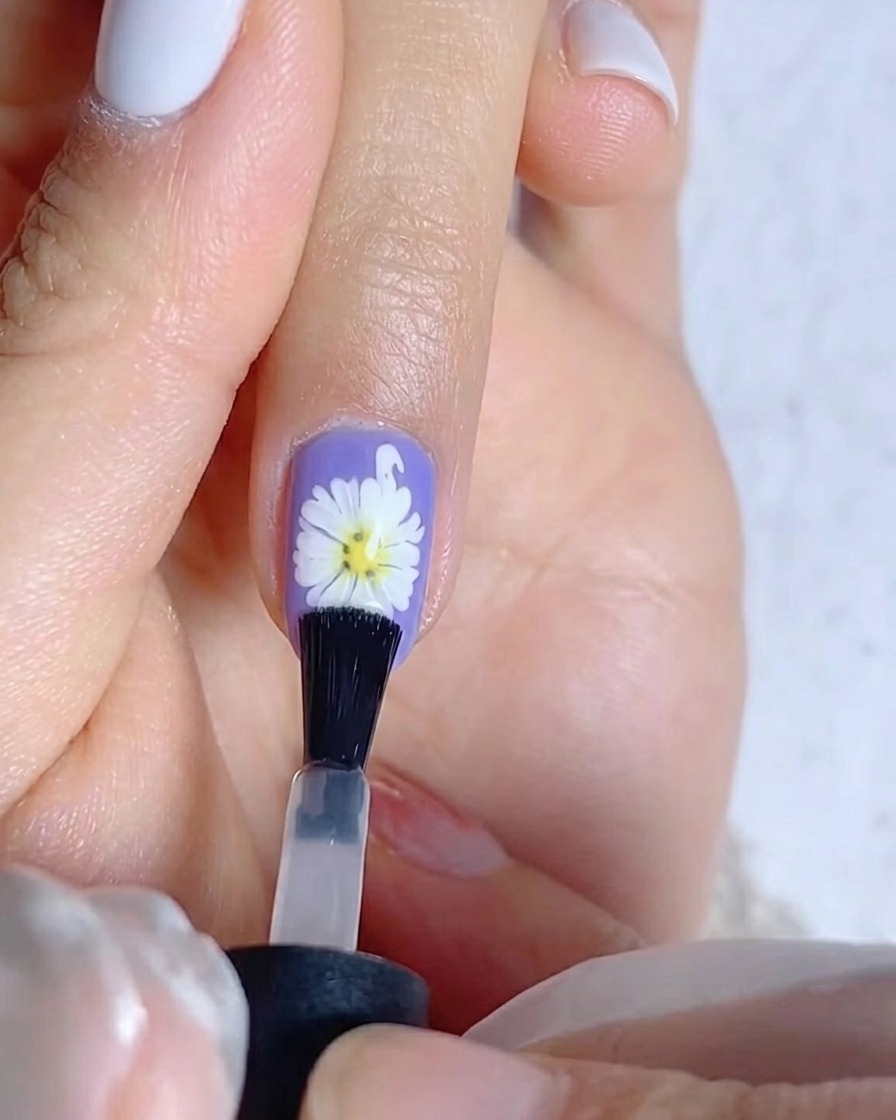 Un pinceau appliquant une couche de gel de finition mat sur un ongle violet présentant un motif de marguerite blanche.