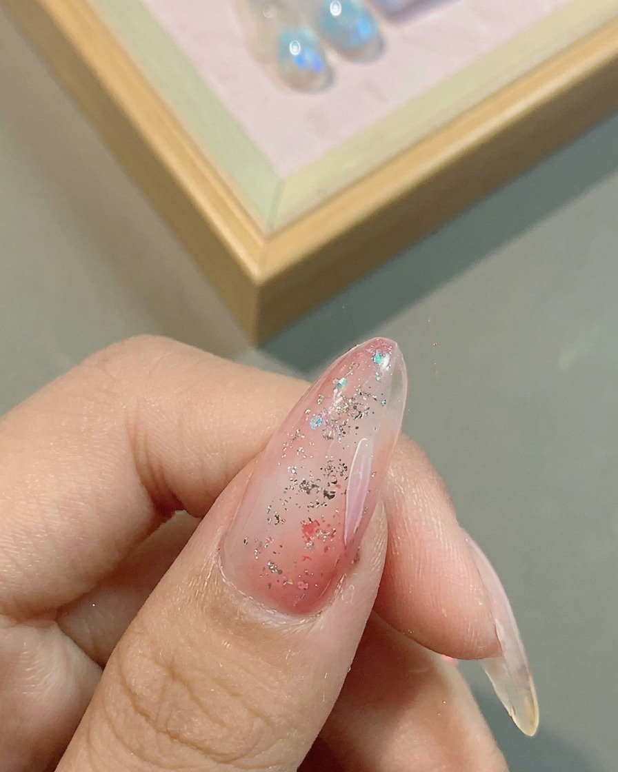 Gros plan sur un pinceau liner fin dessinant des pétales de fleurs blancs délicats sur une base dongle en gel rose et pailleté.