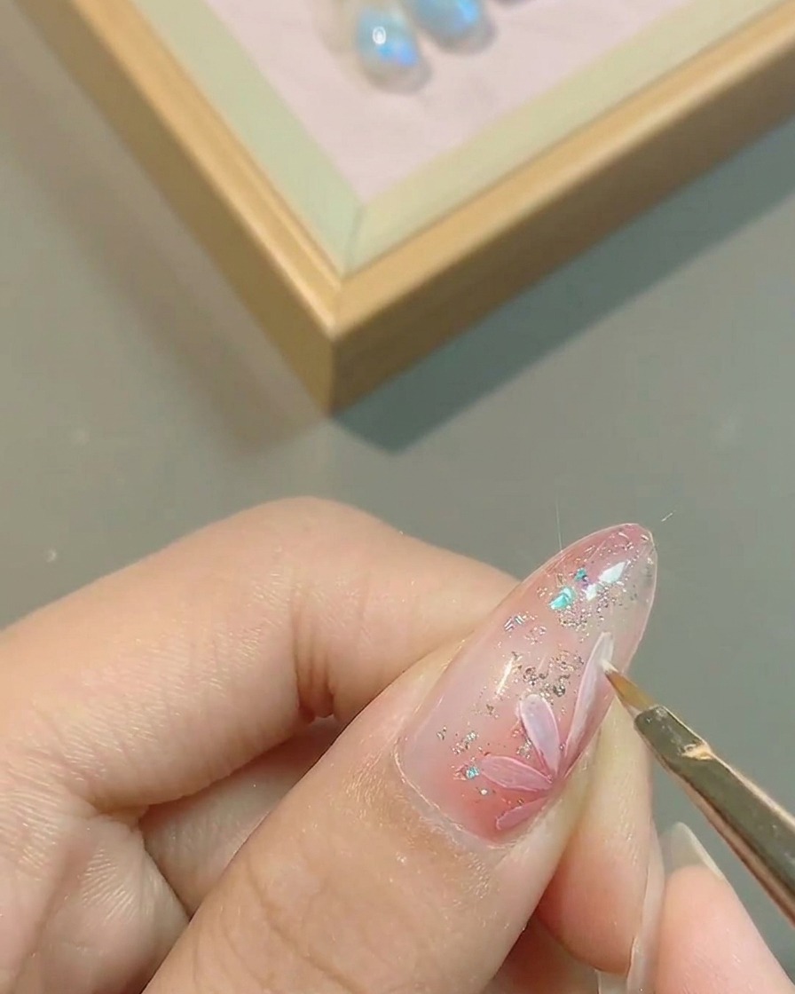 Gros plan dun pinceau traçant précisément les bords des pétales de fleurs blancs peints sur un ongle vernis rose.