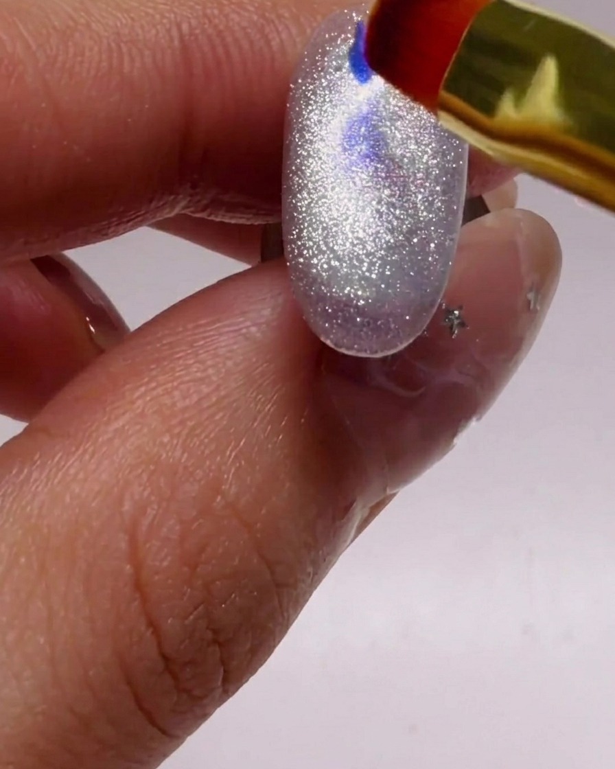 Un petit pinceau déposant des flocons de paillettes bleues et violettes sur un ongle argenté scintillant.