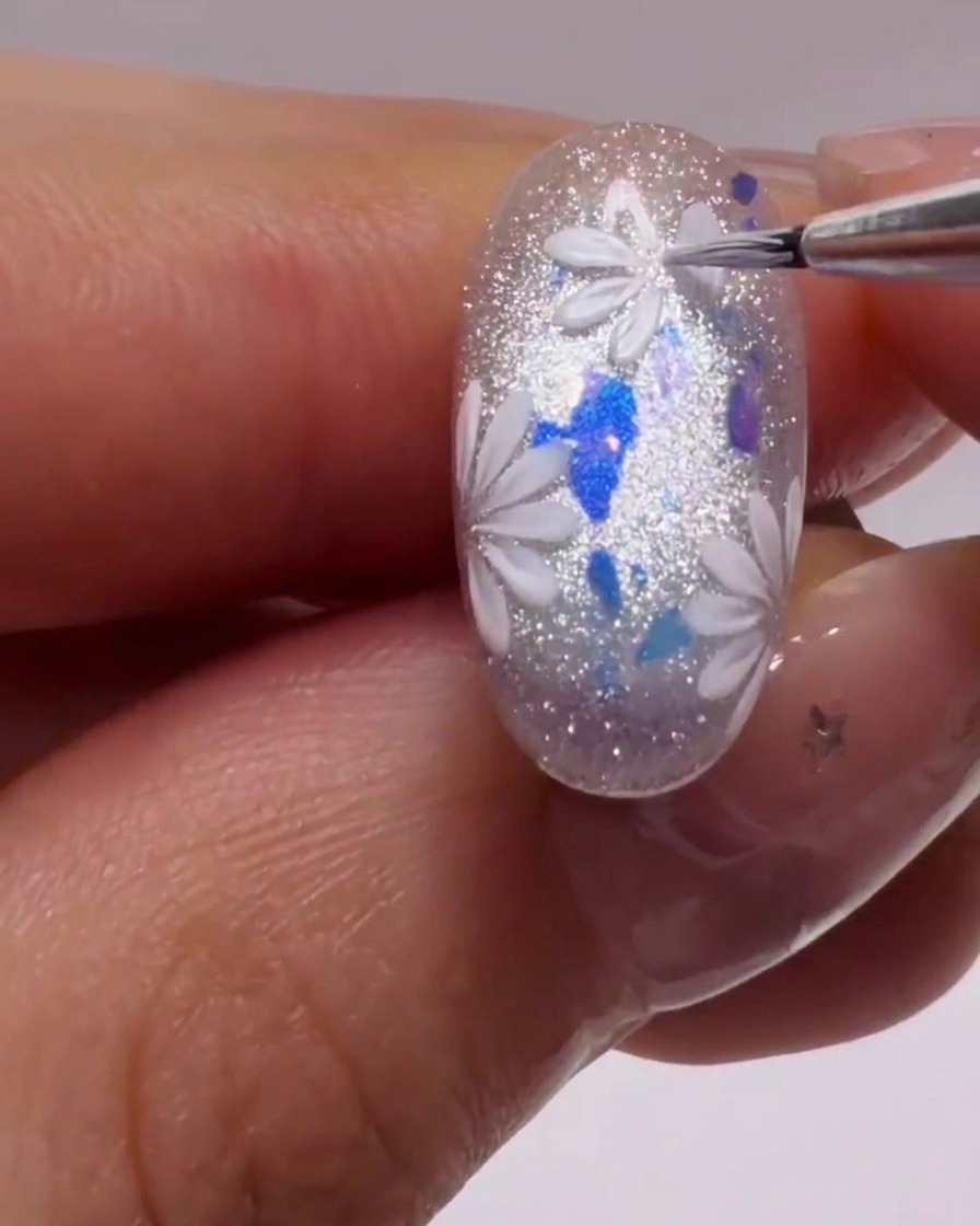 Un pinceau liner fin créant des formes de pétales blancs précis sur un ongle scintillant.