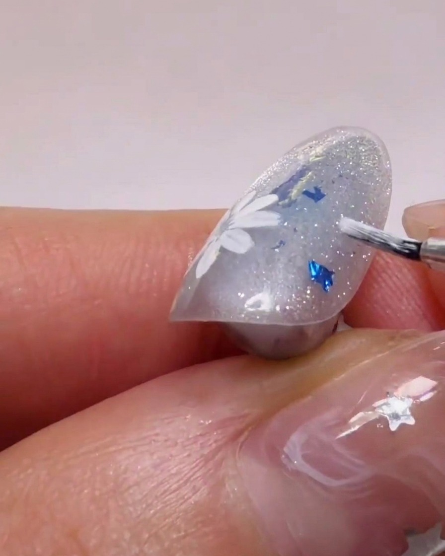 Un pinceau à ongle appliquant du gel blanc pour former des pétales de marguerites sur une base scintillante.