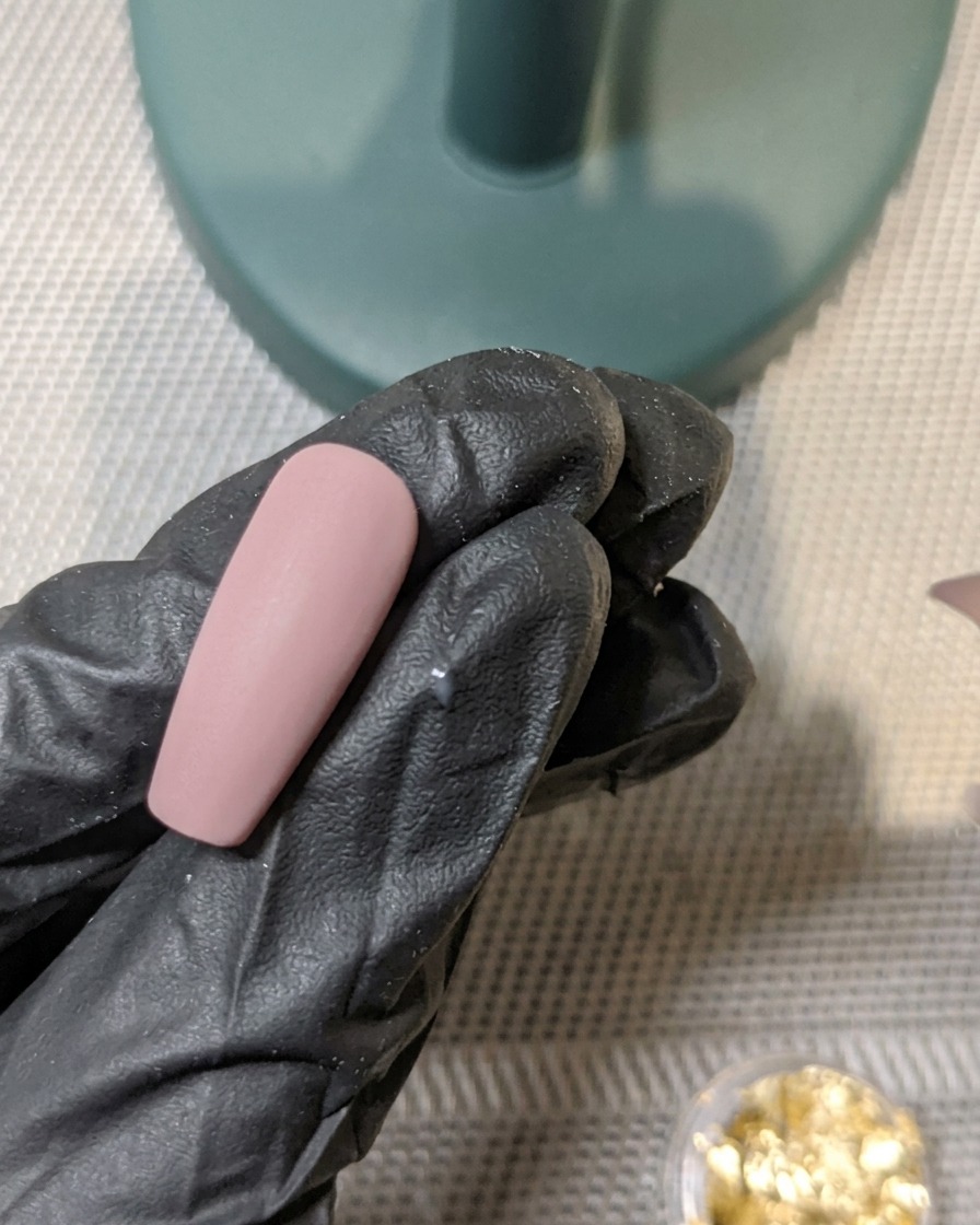 Un pinceau appliquant une couche de top coat mat sur un ongle décoré de feuilles dor incrustées.