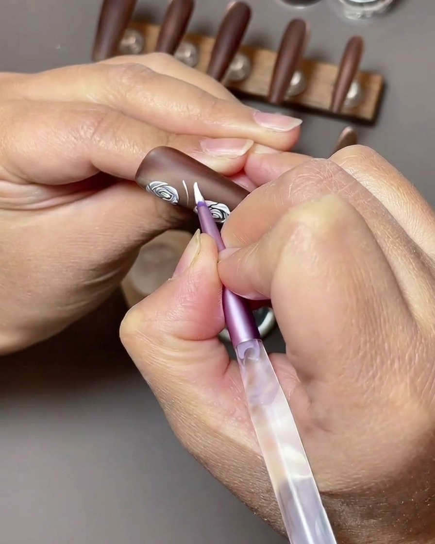 Vue en gros plan montrant du gel nail art blanc utilisé pour peindre des formes de feuilles à côté des roses blanches existantes sur un ongle brun.