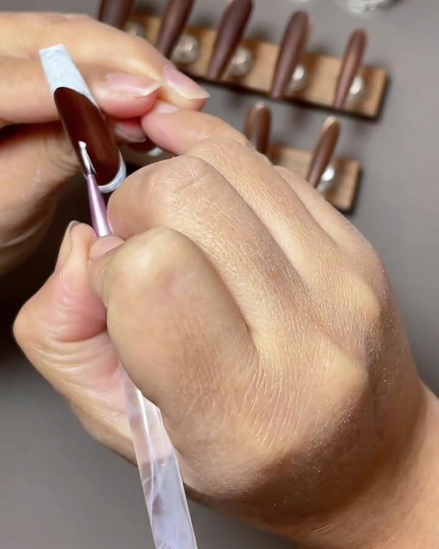 Vue en gros plan dun pinceau nail art fin appliquant une étoile blanche à quatre branches sur un ongle brun foncé avec une French inversée.