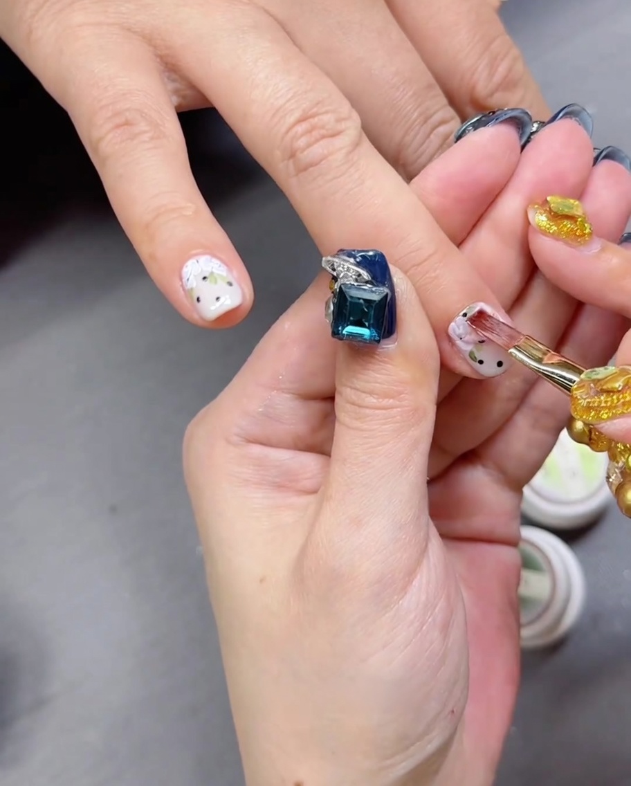 Gros plan sur un pinceau appliquant un top coat transparent brillant sur un ongle à motif de rose et pois.