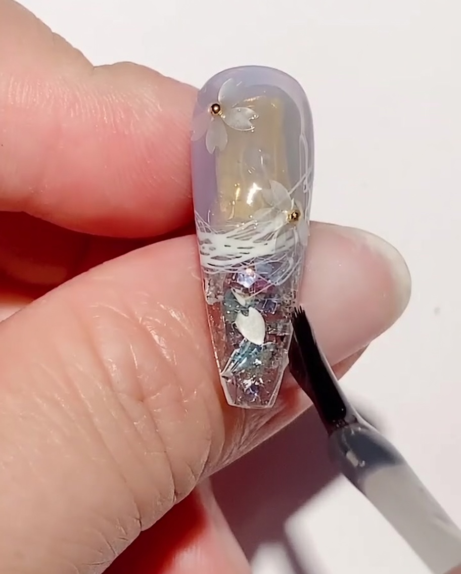 Un pinceau de nail art appliquant une couche de gel de renforcement transparent sur les paillettes et les perles en acier sur un ongle.