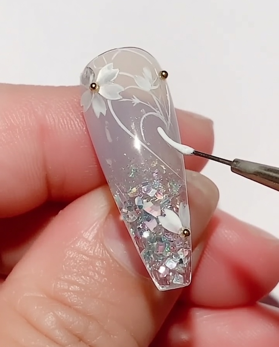 Un pinceau liner fin appliquant du gel couleur blanc pour dessiner une forme de feuille sur un ongle décoré de fleurs de cerisier.