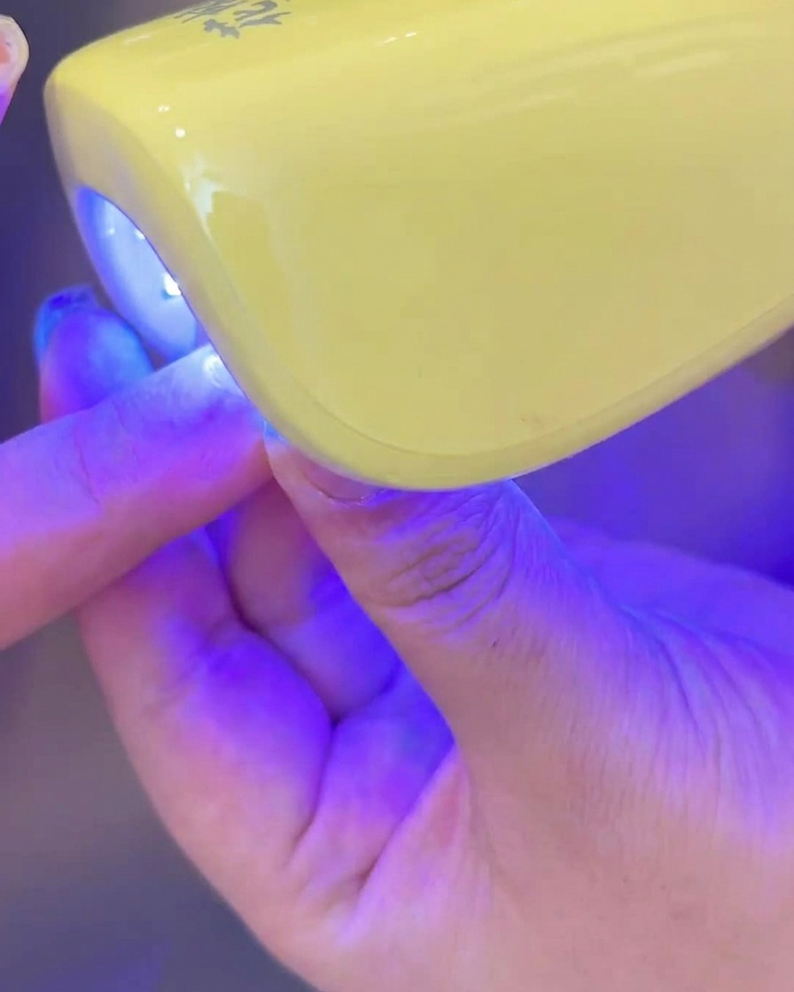 Une main plaçant un ongle avec du gel blanc laiteux appliqué sous une petite lampe UV/LED pour la catalysation.