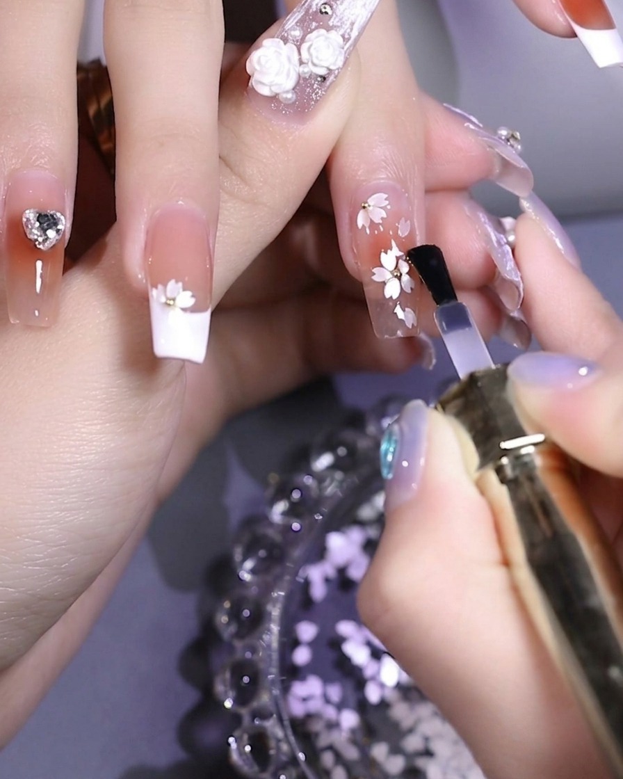 Un pinceau appliquant une finition brillante transparente sur un ongle décoré de fleurs blanches et dune bille métallique centrale.