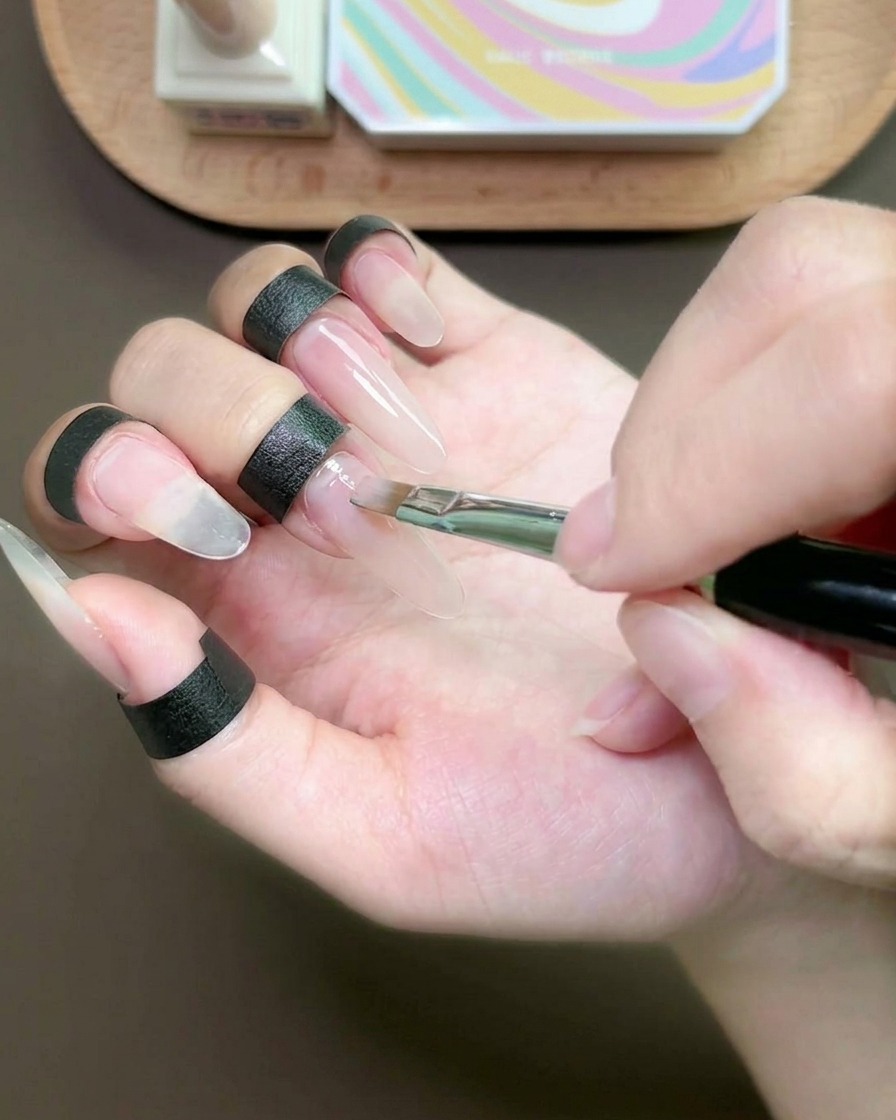 Un pinceau plat appliquant une fine couche de gel nude sur longle du majeur.