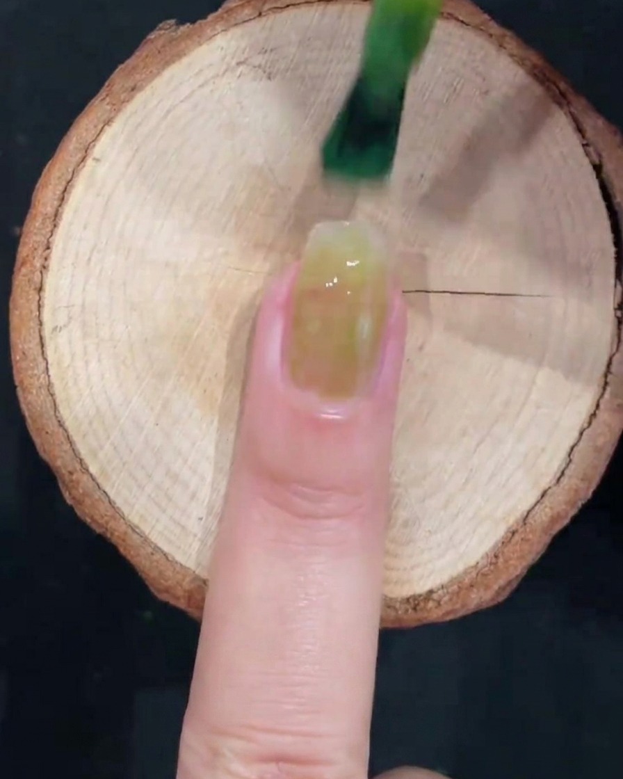 Un pinceau à gel appliquant un vernis vert glacé translucide au centre dun ongle naturel propre sur une surface en bois.
