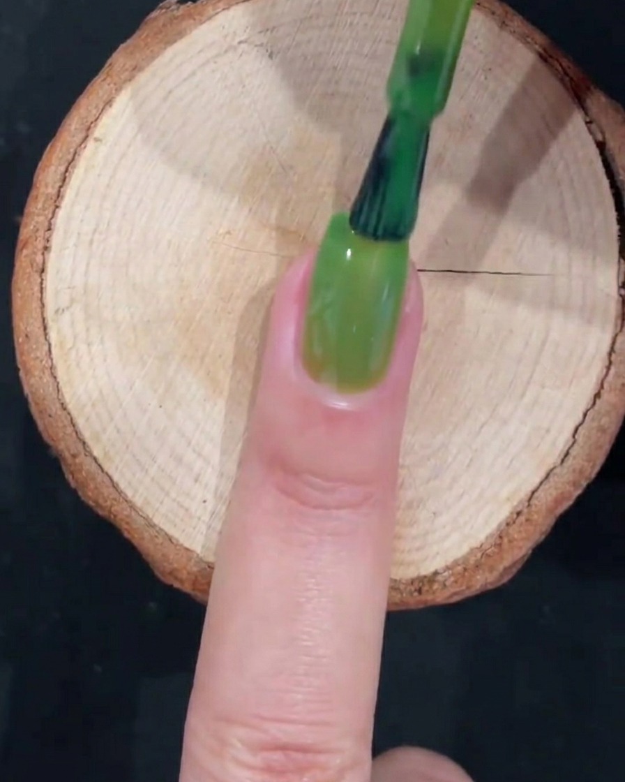 Un pinceau à gel terminant une couche complète de vernis vert translucide avec des mouvements lisses sur la longueur de longle.