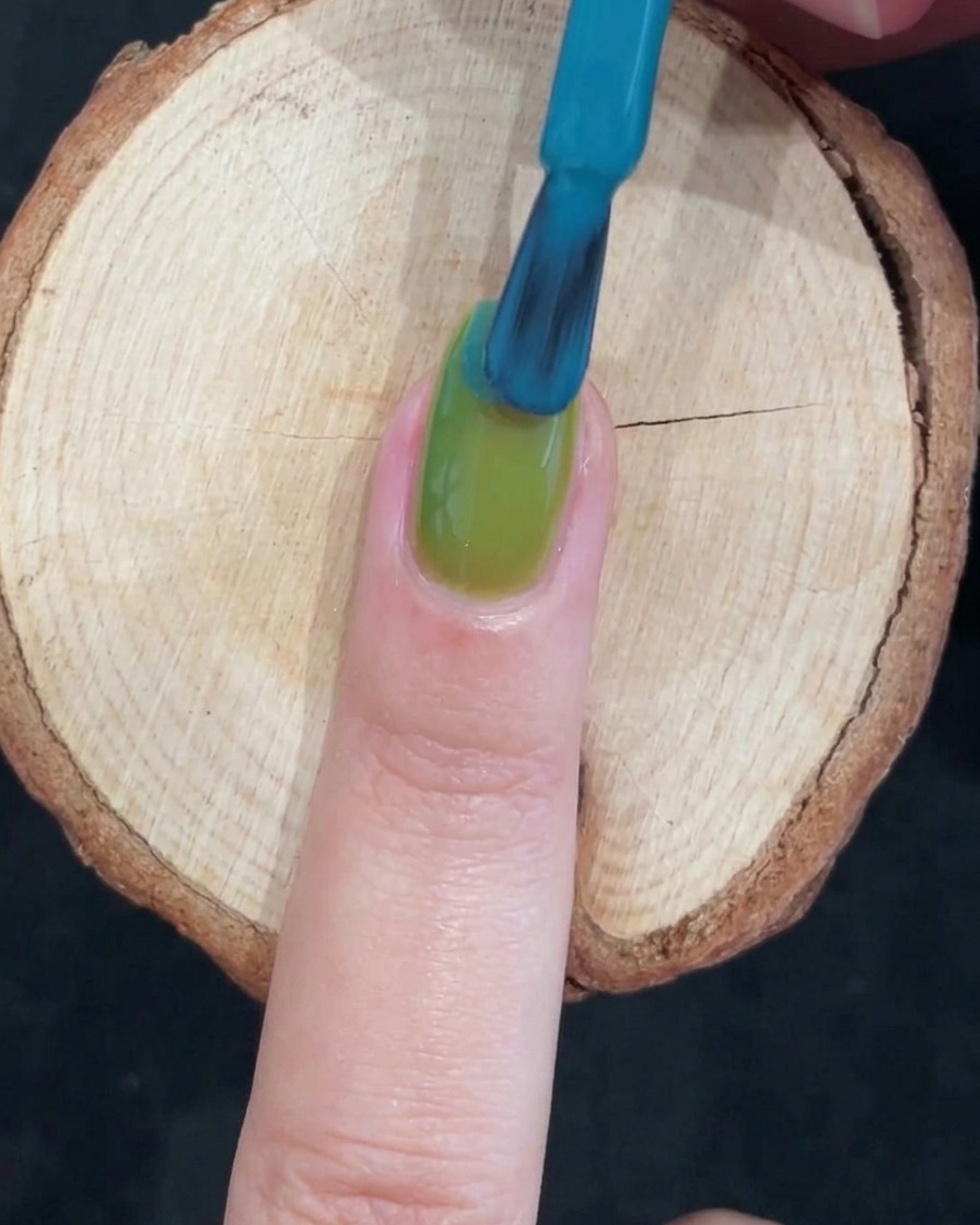 Un pinceau à ongles appliquant un gel bleu vif sur la moitié supérieure dun ongle verni en vert pour créer un dégradé bicolore.