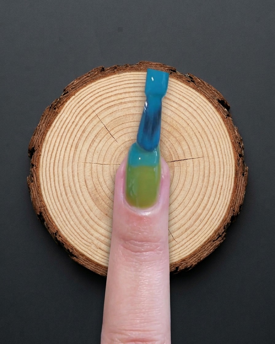 Un pinceau appliquant du gel bleu sur la moitié supérieure dun ongle à base verte.