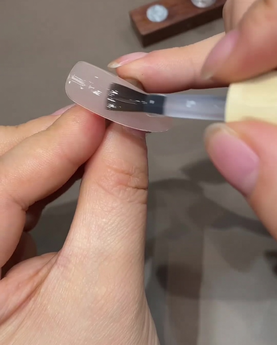 Un pinceau plat pour gel appliquant une fine couche de top coat sur un ongle nude préparé.