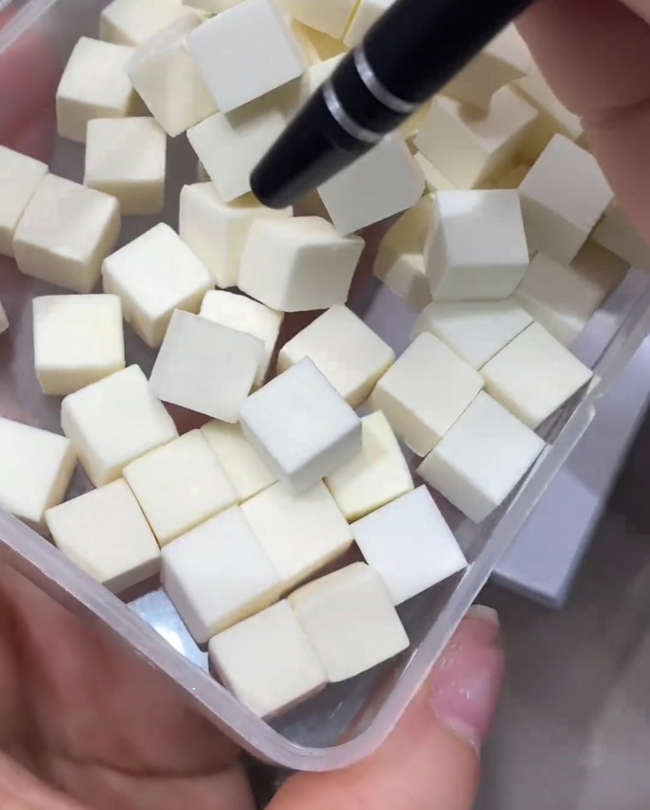Un outil de précision saisissant un petit cube déponge de maquillage blanc dans un récipient en plastique transparent.