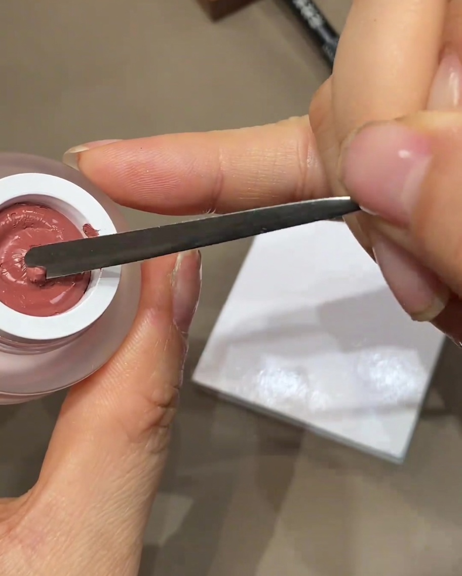 Une spatule métallique déposant une petite goutte de vernis gel coloré sur une palette blanche.