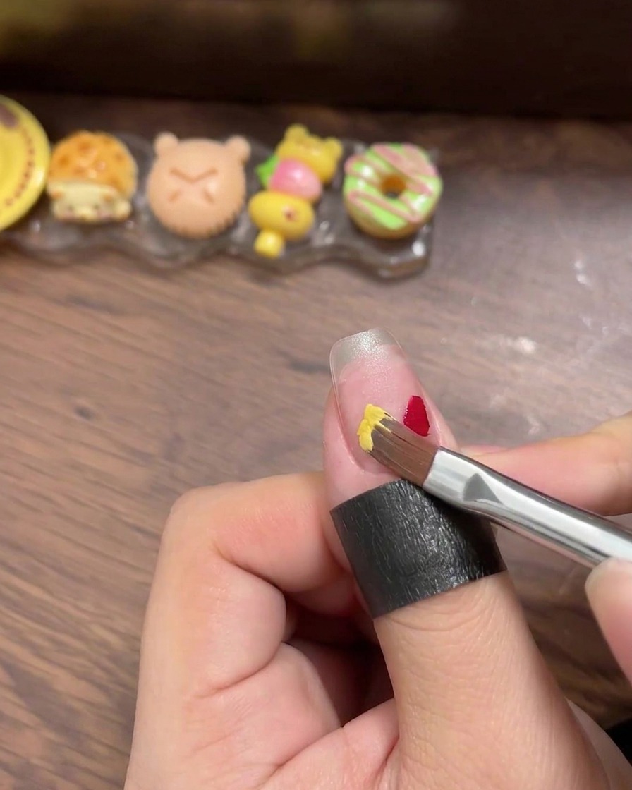 Gros plan sur un pinceau liner appliquant de petits carrés distincts de gel polish rouge, jaune, bleu et menthe sur une capsule transparente.