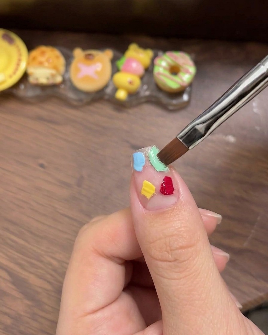 Une éponge à maquillage blanche utilisée pour tapoter et mélanger doucement quatre carrés de gel colorés afin dobtenir un effet ombré lisse sur longle.