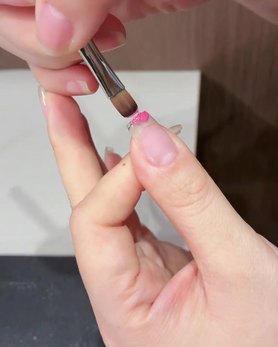 Gros plan sur un pinceau plat appliquant du vernis gel magenta sur le bord libre de longle.