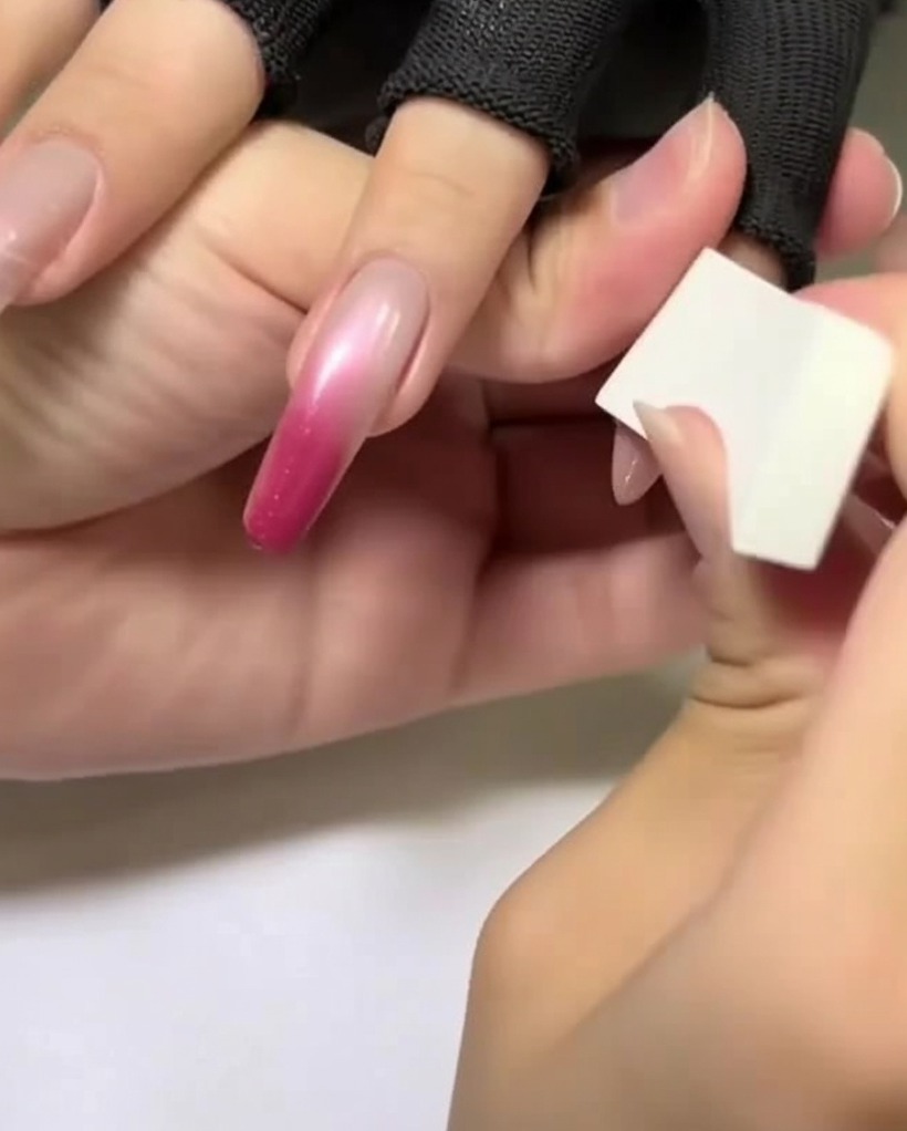 Une main tenant une éponge de maquillage blanche appliquant une teinte rose clair sur la partie supérieure dun ongle en amande.