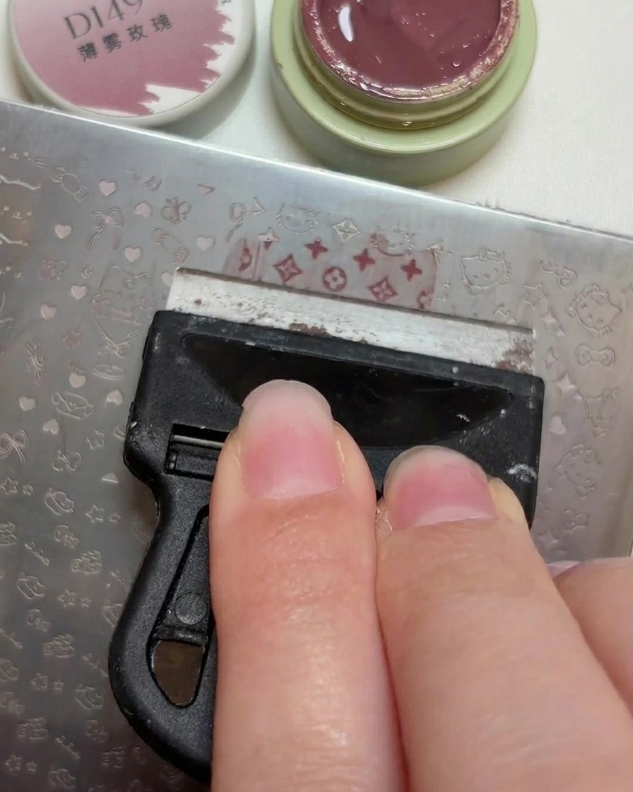 Un racloir en plastique lissant une petite noix de vernis gel vieux rose sur les motifs gravés dune plaque de stamping pour ongles en métalcite: 1.