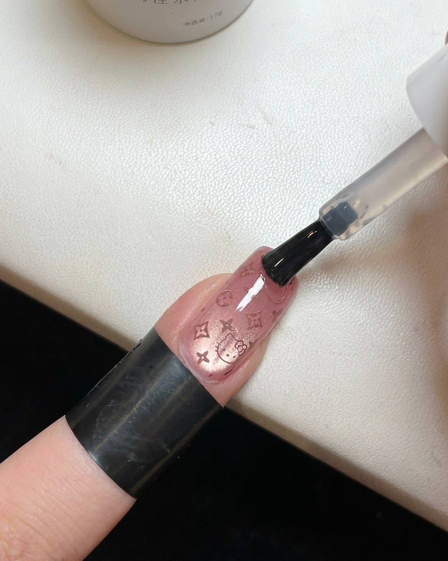 Un pinceau appliquant un top coat transparent et brillant sur un ongle orné dun motif Hello Kitty rosecite: 1.